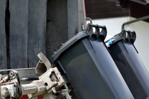 The image shows two black plastic wheelie bins positioned on a paved or concrete surface, attached to a metal rubbish collection vehicle in a driveway or outdoor area. The bins are partially visible, with one in the foreground and the second slightly behind it, both featuring hinged lids at the top and handles on the sides. The vehicle's metallic framework and locking mechanism are clearly visible, indicating it is designed for waste collection. The environment appears to be outdoors, possibly in a residential or commercial setting, with a wooden or metal fence and some architectural elements in the background. The lighting suggests natural daylight, providing clear visibility of the bins' textures and colors, typical of a waste disposal or rubbish removal service operation in Greenwich or nearby areas. This detailed description aligns with services provided by Commercial Waste Greenwich for efficient rubbish removal in the local region.