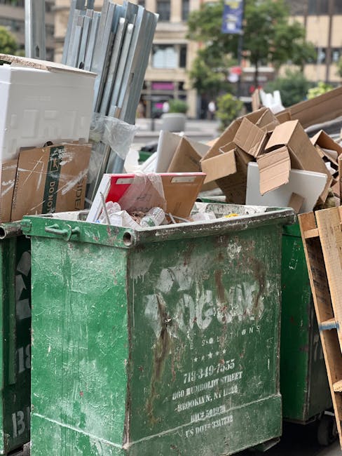 A large, weathered green waste container filled with a mix of cardboard boxes, some flattened and others partially intact, along with white foam pieces and plastic packaging. The container is situated