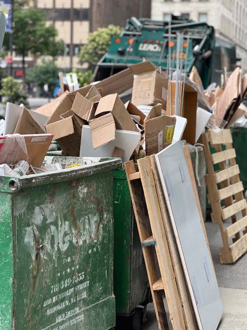 A large, weathered green waste container filled with a mix of cardboard boxes, some flattened and others partially intact, along with white foam pieces and plastic packaging. The container is situated